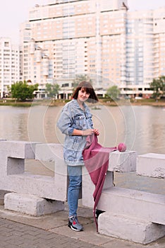 The girl sits by river and knits pink scarf on knitting needles, great weather and the city skyline in the background