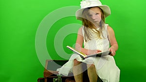 Girl sits on the suitcases and reads the book. Smalll girl on chroma key background. Travelling concept.