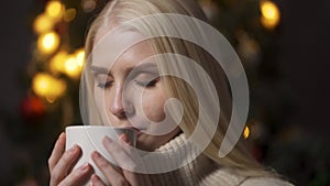 The girl sits under the Christmas tree and drinks cocoa. She holds a glass with both hands