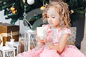 Blond girl in pink dress under the New Year tree and looks at a burning candle