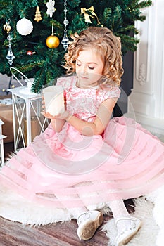 Blond girl in pink dress under the New Year tree and looks at a burning candle