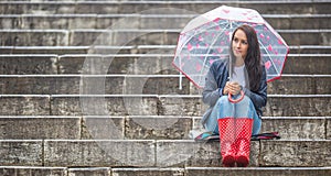 Girl sits waiting on stone stairs, wearing red rain boots and a umbrella with hearts protecting her against the rain