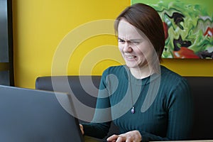 A girl sits, works at a laptop at a table in a cafe on a brown sofa against a yellow wall. On the face is an expression