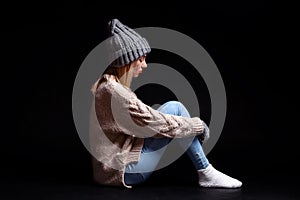 The girl is sitting alone on the floor on a black background of emptiness, hugging her legs with her hands and looking down,