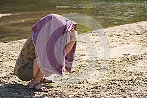 The girl is sitting on the beach with a towel on her head. The girl hid her face from the bright sun under a towel