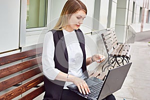 Girl sitting on a bench near a business building with a laptop and bank card.