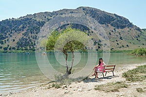 A girl is sitting on a bench near a tree on lake kurnas. Island of Crete.