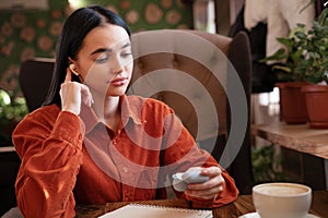 Girl sitting with earphone in ear holding charging case for wireless headphones while relaxing in cafe