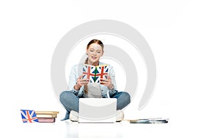 Girl sitting on floor with laptop, copybooks and uk flag while reading book