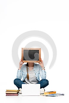 Girl sitting on floor with obscure face near laptop, books and copybooks and holding chalkboard