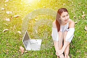 A girl sitting on the grass and labtop near her. A glasses is on the labtop.