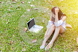 A girl sitting on the grass and labtop near her. A glasses is on the labtop.