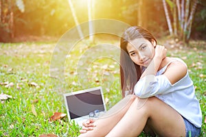 A girl sitting on the grass and labtop near her. A glasses is on the labtop.