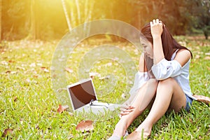 A girl sitting on the grass and labtop near her. A glasses is on the labtop.