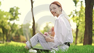 Girl sitting in the grass writing something in notes and working with a laptop and looking at camera