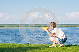 The girl is sitting on the green grass and playing with a water pistol, against the blue sky and the lake, there is a place for an