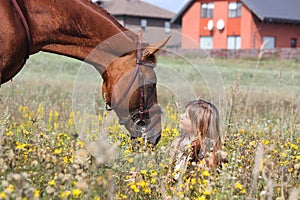 Girl sitting on the ground and chestnut horse standing near