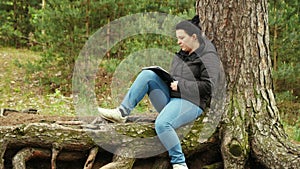 Girl sitting on large tree roots and writing something in a notebook.