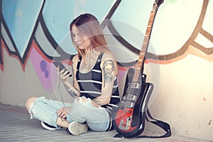 Girl sitting near graffiti wall with a guitar listening music.