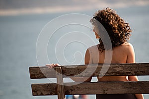 A girl sitting over her shoulder on a wooden bench looks at a mountain lake.