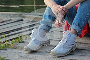 Girl sitting on a river beach on a wooden pier. Closeup of teenage legs in modern and trendy white sneakers and rolled up jeans.