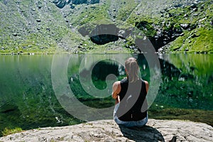 A girl sitting on a rock, looking at a spectacular view of the mountain reflecting in the lake.