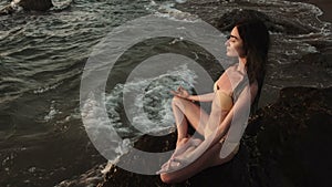 Girl sitting on rock by sea, meditating in lotus yoga pose, concentrating.