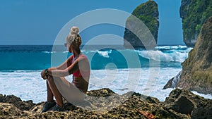 Girl sitting on the Rock and watching Huge Waves hitting Tembeling Coastline at Nusa Penida Island, Bali Indonesia