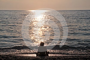 Girl sitting on sandy beach next to sea and enjoys watching sunset, reflection of sun rays over sea. Silhouette of woman by ocean.