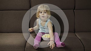 Girl sitting on sofa and eating corn puffs. Child smiling and taste puffcorns
