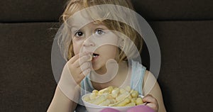 Girl sitting on sofa and eating corn puffs. Child taste puffcorns