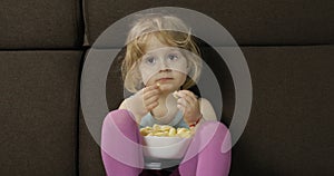 Girl sitting on sofa and eating corn puffs. Child taste puffcorns