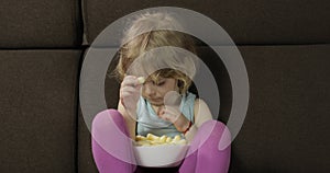 Girl sitting on sofa and eating corn puffs. Child watching tv, taste puffcorns