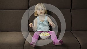 Girl sitting on sofa and eating corn puffs. Child watching tv, taste puffcorns