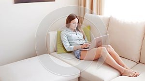 Girl sitting in a sofa and typing something on her laptop. Young woman using the laptop at the sun set.