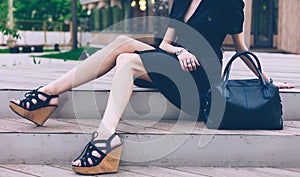 Girl sitting on the stairs with a big black super fashionable handbags in a dress and High Wedge Sandals on a warm summer evening