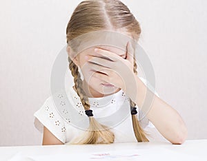 Girl is sitting at the table and covers her face with her hands.