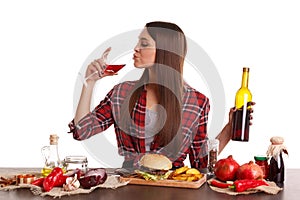A girl sitting at a table with food, drinking red wine from a glass and holding a bottle of red wine.