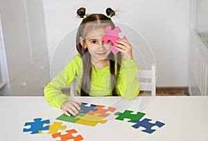 Girl is sitting at a table with puzzles and closes eye with one detail