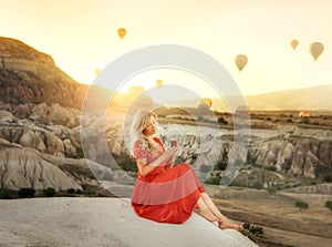 A girl sitting on the top of a cliff with a glass of Turkish tea at dawn with a view of the mountains of Cappadocia and balloons i