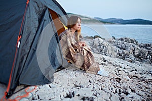 Girl sitting under tents, wrapped in plaid, holding cup of fragrant tea.