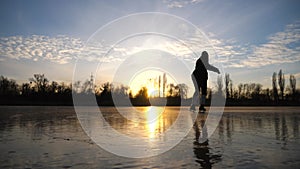 Girl skating on frozen river during beautiful sunset. Young woman shod in figure skates sliding on ice. Shining sun is