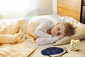 A girl is sleeping in bed, early morning, the sun is shining in the window and next to the alarm clock. Healthy sleep.