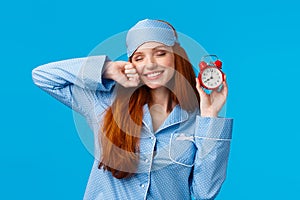 Girl slept well, wake up energized, sleepy stretching with closed eyes and lovely smile, holding red cute clock, set up