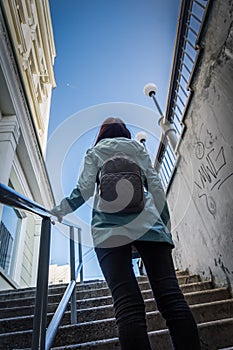 A girl with a small city backpack walks out of the subway, out of the dark, into the light and clings to the handrail.