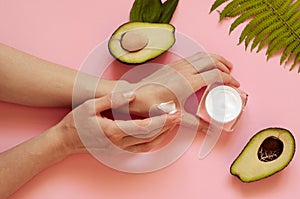 Girl smears cream hands. Two halves of avocado with leaves, a jar of cream and a leaf of fern on a pink background. Natural Organi