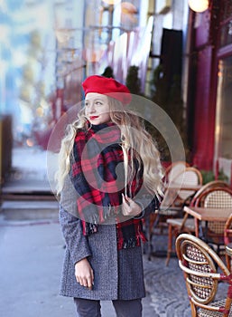 Girl in a gray coat and a red beret with long curls stands thoughtfully near a cafe on a background of a red facade