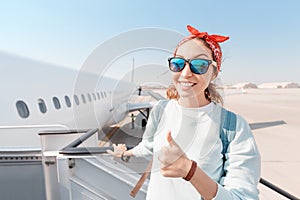 Girl smiles and gives a thumbs up while boarding a large white plane at the airport. Concept of travel and victory over