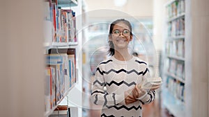 A girl is smiling and holding a stack of books in a library