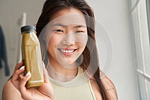 Girl smiling toothy and looking straight while showing bottle with smoothie